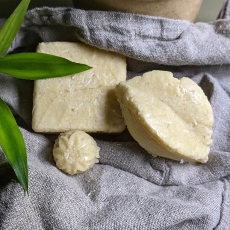 Three Lassëa solid shampoo bars, one flower-shaped, on gray linen beside green leaves